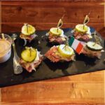 Zesty Reuben Bites - A flavorful stack of cucumber and pickles, layered with corned beef, sauerkraut, Swiss, and Dubliner cheddar cheeses, served with a side of Russian dressing for dipping, plated on a slate serving board.